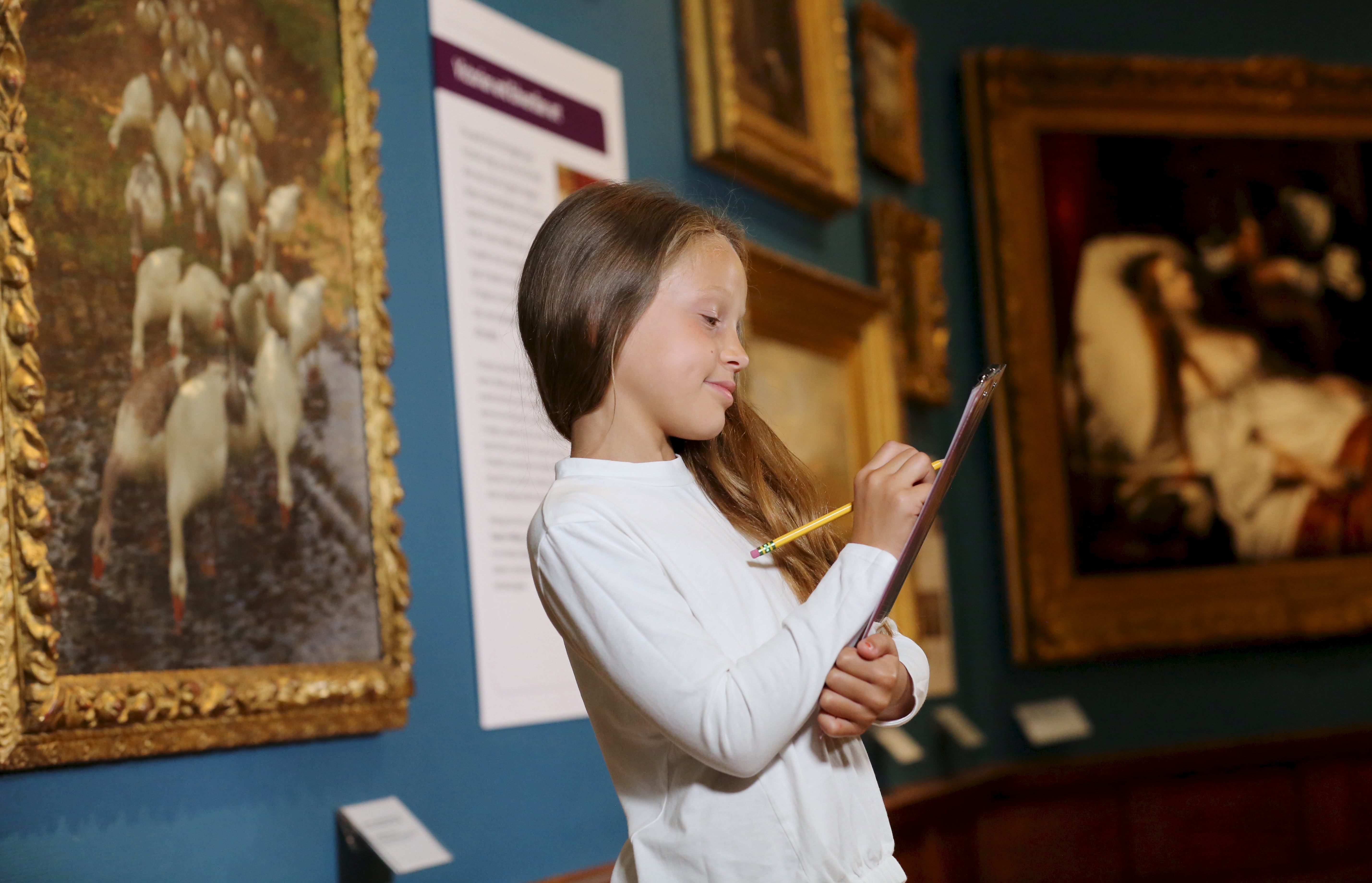 A girl with a clipboard in the gallery, which has blue walls and a painting of geese on the wall